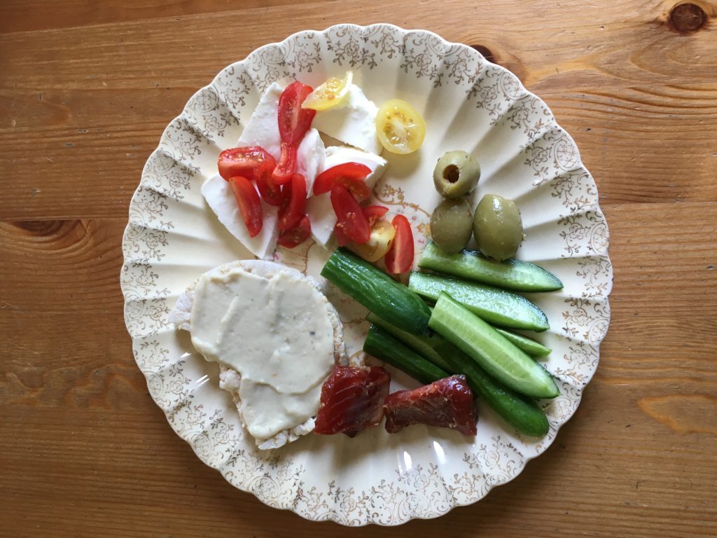 A white plate on a wooden table. The plate contains red tomatoes, white mozzarella cheese, olives, cucumbers and salmon chunks.