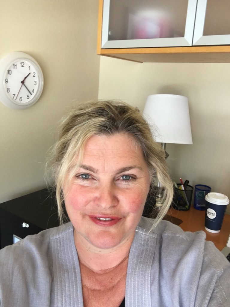 Elizabeth in an office pictured smiling  standing in front of a desk. On the desk is a take out coffee cup, a lamp and some pens in a holder. There is a clock and cabinet on the wall behind her.