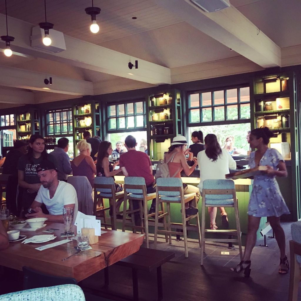 Photo shows a picture of a bar where a number of people are seated on high chairs with their backs to the camera. They are facing a bartender behind which are two large open windows with other restaurant patrons seated on the opposite bar looking in towards the restaurant. In front of the closest bar seats is a wooden table where a diner is seated. He is wearing a white t-shirt and facing the camera. The ceiling is painted white, the window trim is green and a waitress in a short blue dress is walking by carrying menus.