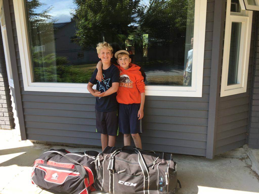 Two boys one taller than the other standing in front of the window of their house. In front of them are two filled hockey bags. they are smiling.