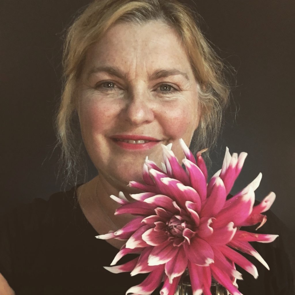 This is a picture of a smiling Elizabeth wearing a dark shirt against a dark backdrop. She is holding one large pink dahlia flower that has white tips. 