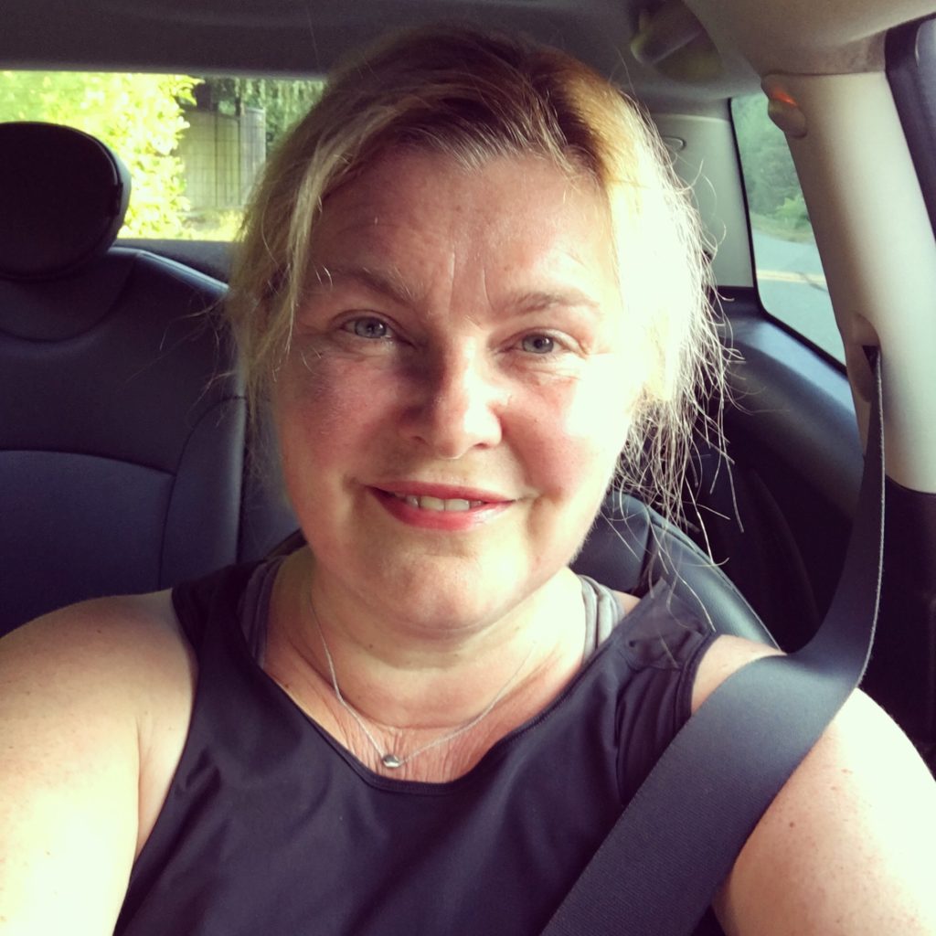 A picture of Elizabeth sitting in her car with her seatbelt on. She is smiling and sweaty from her morning gym workout. She is wearing a black top.