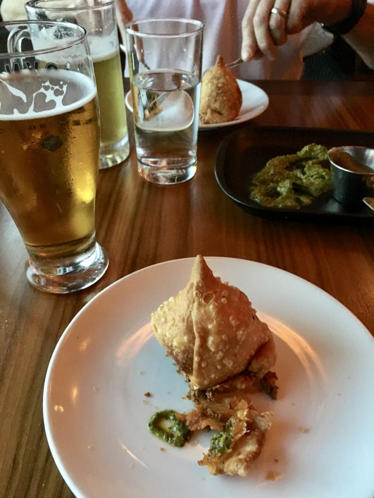 A deep fried samosa on a white plate, with a piece broken off and on the plate is some green sauce