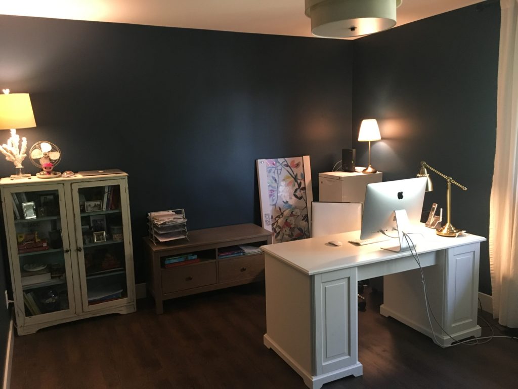 Elizabeth's newly decluttered office. The walls are dark blue and the ceiling and drapes are white. The floor is dark brown. To the left is a small bookshelf with a lamp and old fan on top. Her desk has a silver computer monitor on top and a brass light. behind the desk to the left is a low grey table with books and files in a tray. To the left is a picture of flowers waiting to be hung and next to it a filing cabinet. The surface of the white desk is empty and the office looks neat.