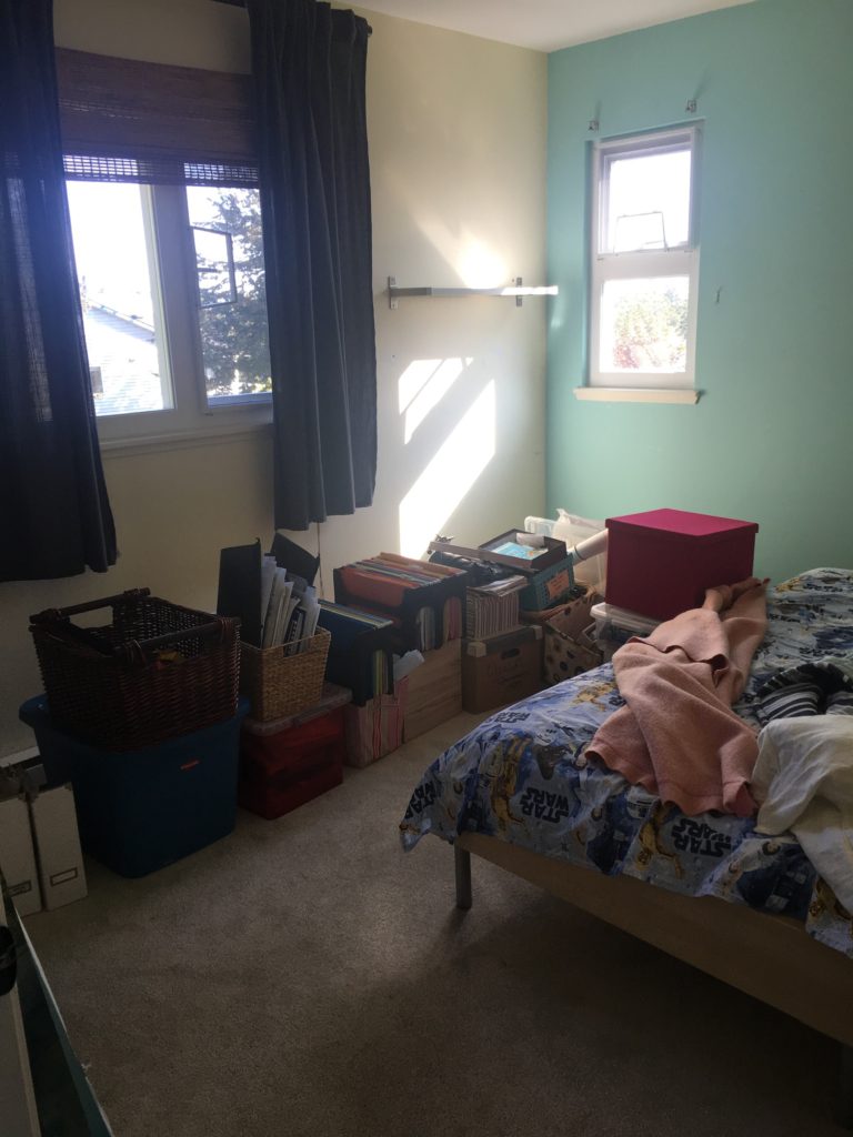 A bedroom, the wall to the left is pale yellow the wall to the right is a light turquoise. The large window has a rattan blind and dark grey drapes and the small window has no window covering. The sun is shining through the small window onto a pile of boxes sitting on the beige carpet. There is a single bed at the front that has unmade Star Wars sheets on it and a pink wool blanket.