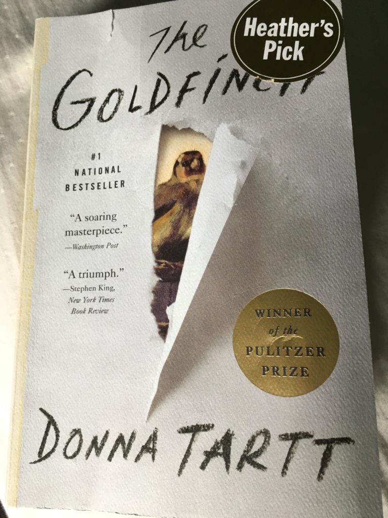 A picture of a book, the cover is white with what looks like a tear in the paper in the middle of the cover, underneath is a brownish yellowish gold finch peering out through the tear. The cover reads 'The Goldfinch' by Donna Tartt winner of the Pulitzer Prize. 