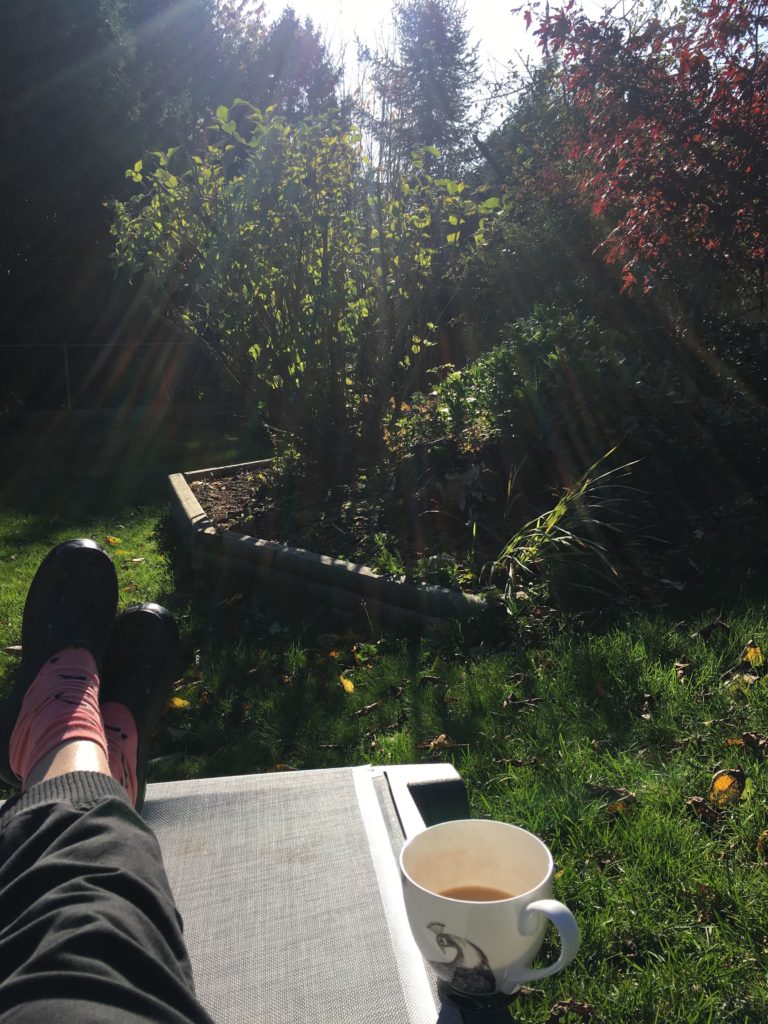 Elizabeth is lying on a chaise lounge chair in her backyard but only her legs are visible in the photo. Beside her is a tea cup filled half way with tea. She is wearing black pants, orange socks and black slip on crocs. The sun is shining down on the trees and shrubs in front of her. 